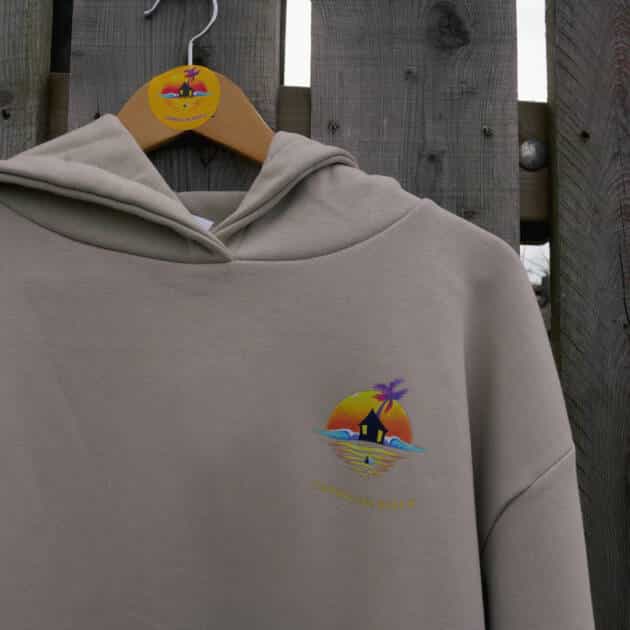 A beige Shack Brand Curracloe Beach Adults' Hoodie hangs on a wooden hanger before a fence, displaying small embroidery of a sunset, palm tree, hut, waves, and the words "CORALLOUS BEACH.