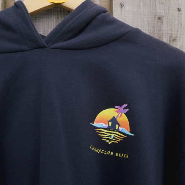 Shack Brand Curracloe Beach Women's Hoodie in navy hangs on a wooden hanger. Features a colourful sunset, palm tree, waves, and hut logo on the front and the round hanger tag above.