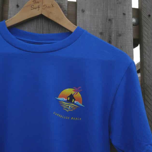 A blue Shack Brand Curracloe Beach Kids T-shirt hangs on a wooden hanger labelled "The Surf Shack," featuring a sunset, hut, palm tree, and waves graphic with the text "Curracloe Beach" underneath.