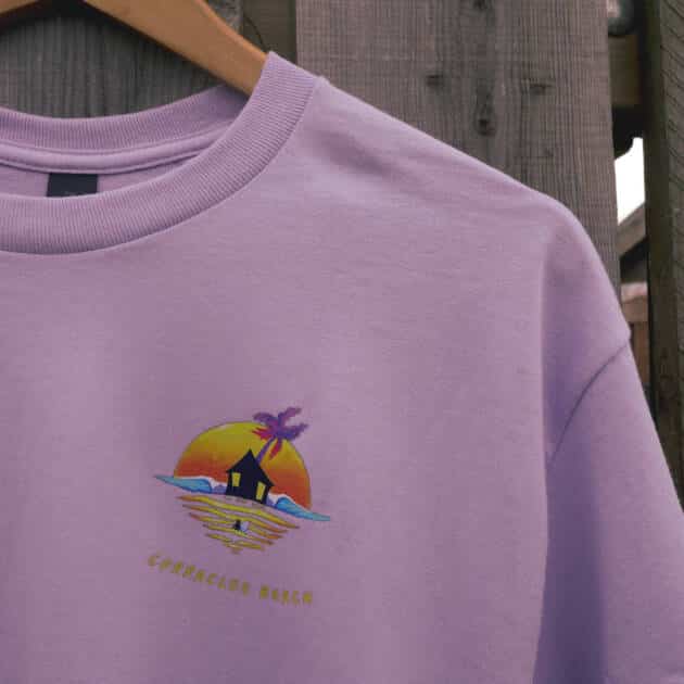 A Shack Brand Curracloe Beach Adult T-Shirt in lavender hangs on a wooden hanger, featuring a vibrant hut, waves, palm tree, sunset graphic, and "CORRALCLOO BEACH" text beneath the design.
