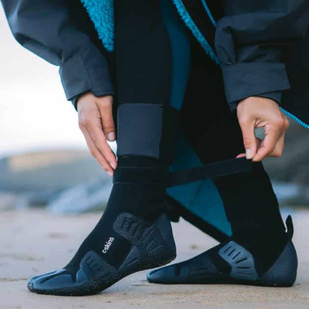 A person stands on a sandy beach in black Cskins Session 5mm Split Toe Boots and a black wetsuit, adjusting the Velcro strap on one boot. A blue fleece-lined jacket is also visible, with sand and rocks blurred in the background.