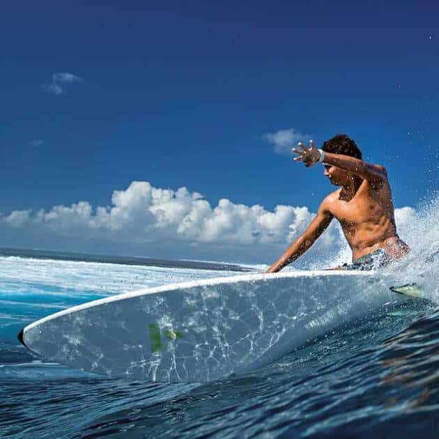 A surfer rides a wave on a sunny day, leaning forwards on a large white Tahe (BIC) surfboard with one arm extended, creating a splash of water under a clear blue sky with some clouds.