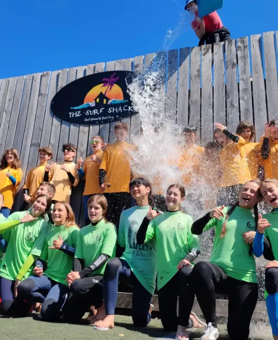 A group of people in green and yellow shirts pose and smile in front of a wooden wall with "THE SURF SHACK" sign, as water splashes down from above, capturing a fun and energetic moment outdoors.
