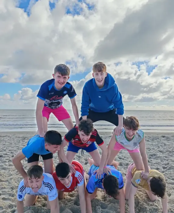 A group of boys on a sandy beach form a three-tier human pyramid, smiling and laughing with the sea and a cloudy sky in the background.