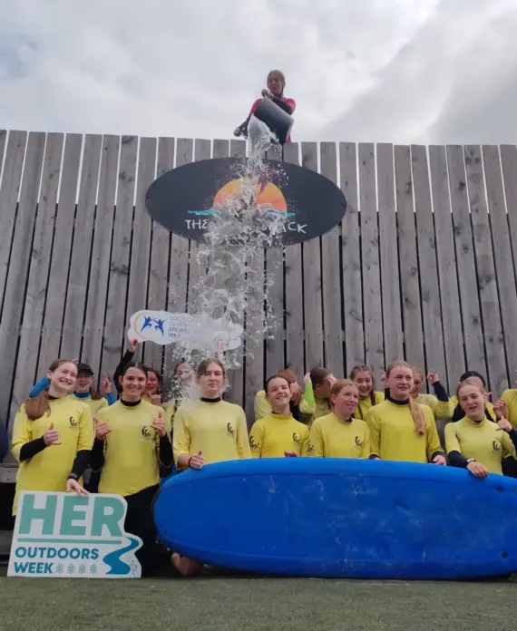 A group of girls in yellow shirts pose with a surfboard and signs reading "HER Outdoors Week" and "HERMOVES" as water is poured on a person above them, standing on a wooden platform.