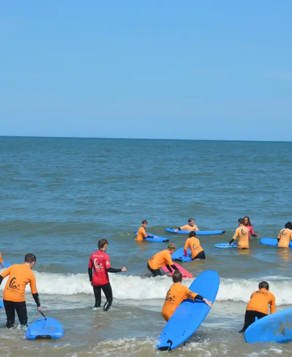 A group of people in orange and red shirts with blue surfboards stand and kneel in shallow sea water, learning to surf under a clear blue sky.