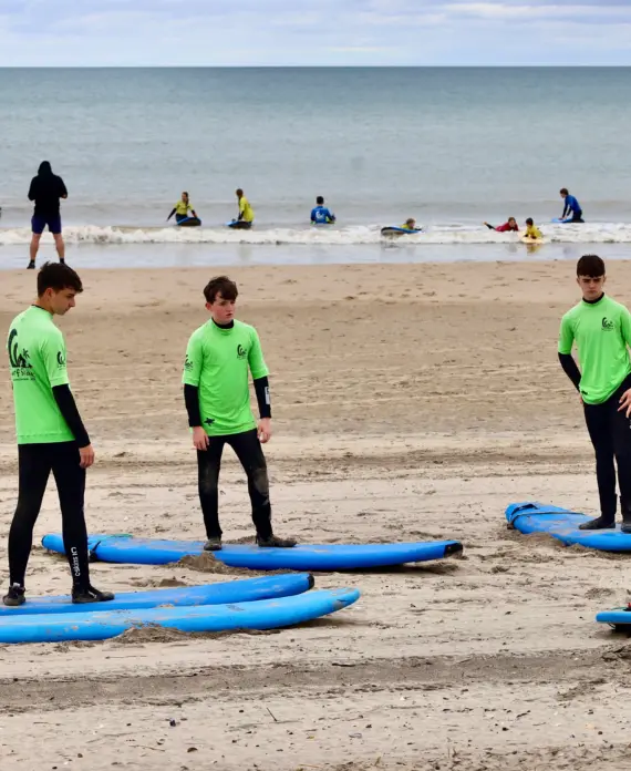 A group of four people in green shirts stand on the beach around blue surfboards, listening to an instructor in a red shirt. More people surf and play near the shore and in the water in the background.