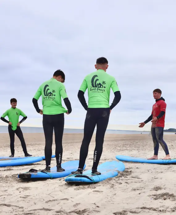 A surf instructor explains techniques to four students standing beside blue surfboards on a sandy beach. The students wear green shirts and wetsuits, while the instructor wears a red shirt. The sky is overcast.