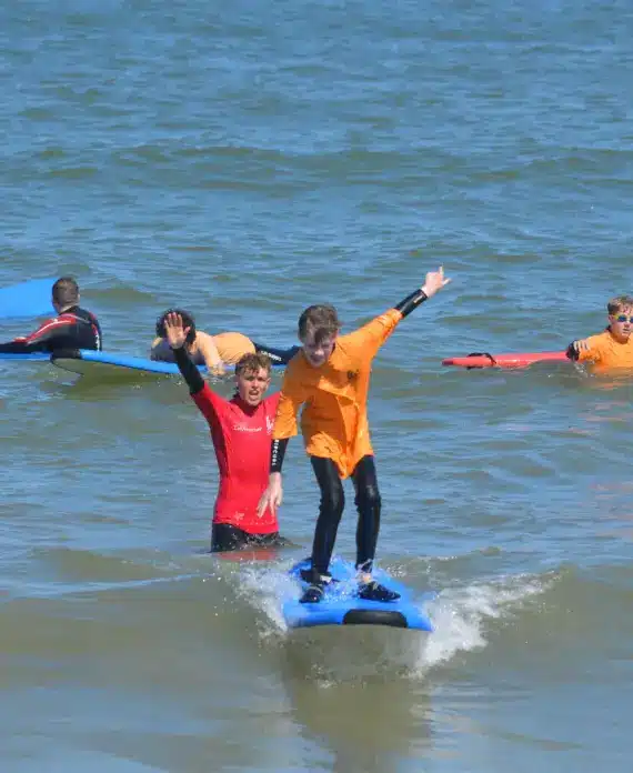 A child in an orange shirt stands on a blue surfboard, balancing with arms outstretched, while an instructor in red supports him. Other children in orange shirts paddle on surfboards in the background.