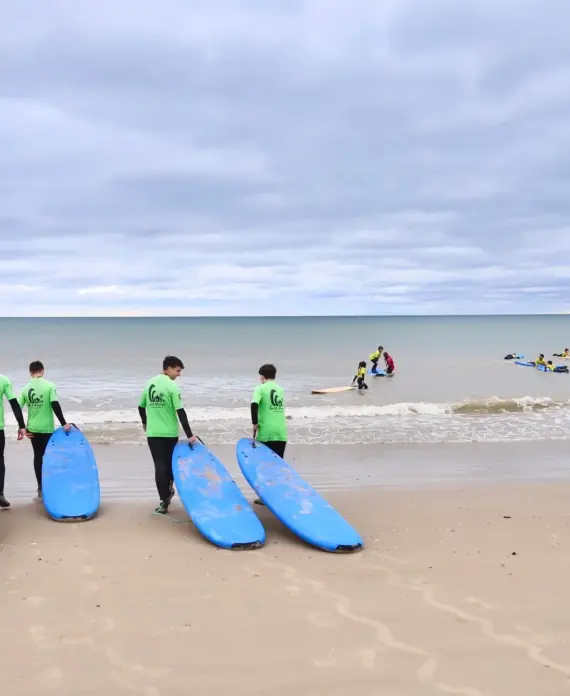 A group of people in green shirts carrying blue surfboards walk towards the sea, whilst others in the water are surfing and paddling under a cloudy sky at a sandy beach.
