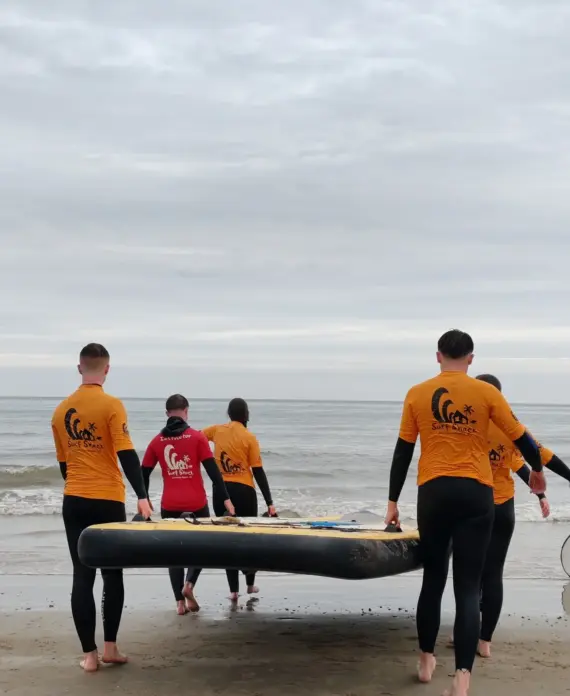 Five people in wetsuits, four in orange and one in red, carry a large surfboard towards the sea on a cloudy day. They are walking barefoot on the sand, heading towards calm water under an overcast sky.