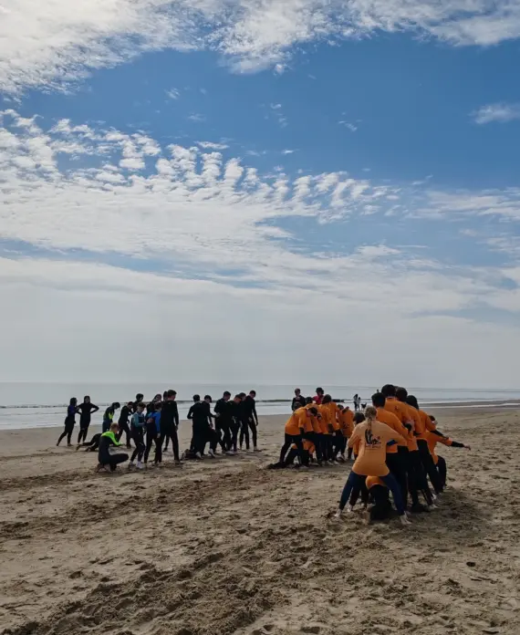 Groups of people wearing wetsuits and orange shirts participate in an outdoor team activity on a sandy beach under a partly cloudy sky.