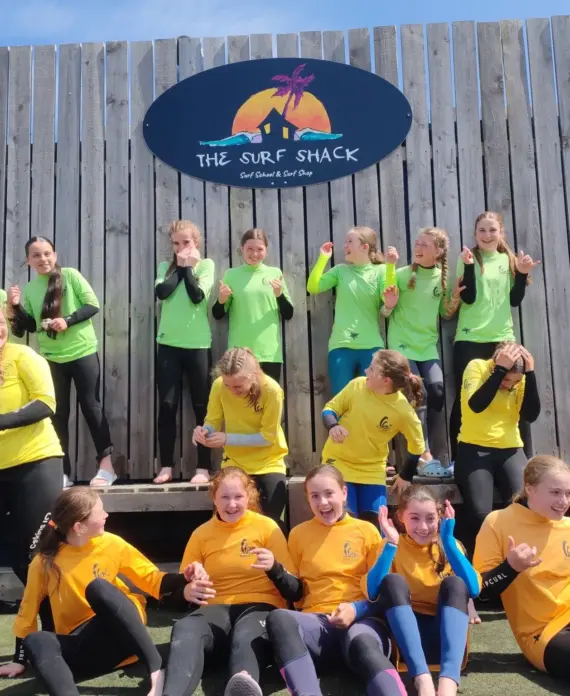 A group of girls in yellow and green shirts and wetsuits pose and laugh in front of a wooden wall with a sign reading "The Surf Shack." The sky is blue and everyone looks happy and excited.