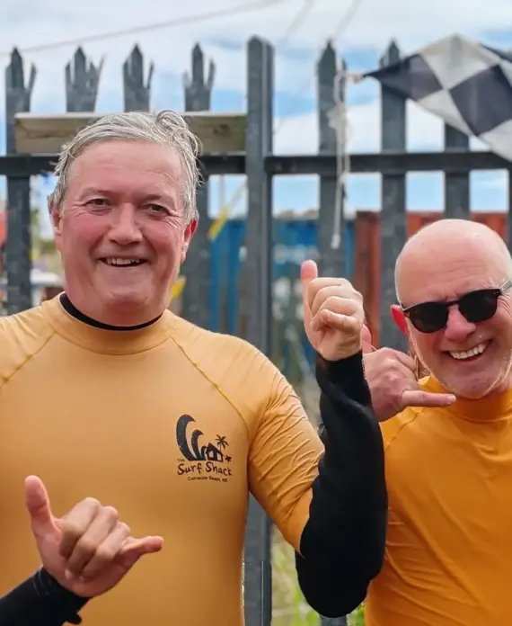 Four people in wetsuits smile and make shaka hand signs in front of a fence and a chequered flag. Three wear yellow tops and one wears red. The background shows blue sky and outdoor equipment.