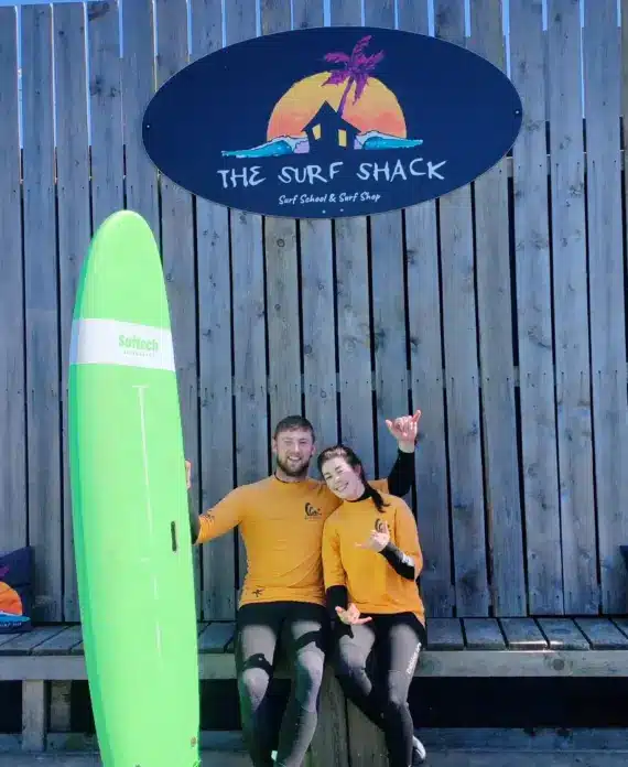 Two people in yellow surf shirts sit on a bench under a wooden sign reading “The Surf Shack” with a palm tree and sunset logo. A green surfboard stands upright next to them. They both smile and pose for the camera.