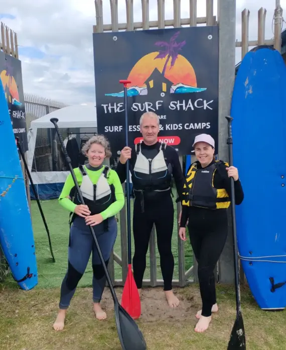 Three people in wetsuits stand smiling with paddles in front of a sign that reads "The Surf Shack." Two blue surfboards are propped up nearby, with a building and greenery in the background.