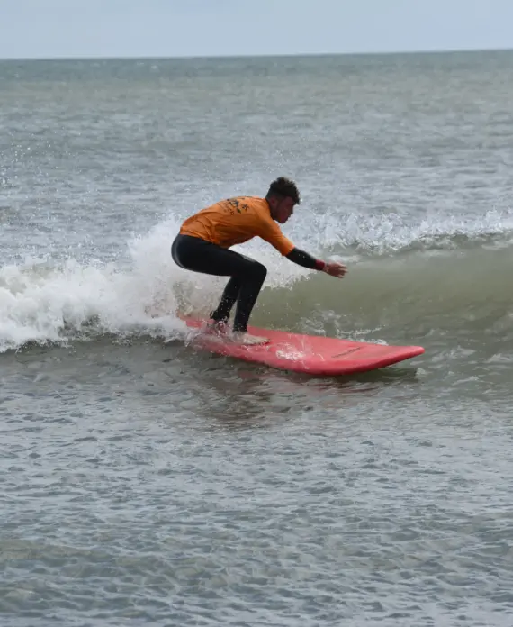 A person wearing a long-sleeved orange top and black trousers is surfing on a small wave using a red surfboard, with another person in the water in the background. The sea appears calm and overcast.
