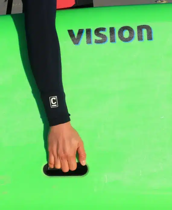 A person in a wetsuit holds a bright green surfboard with the word "VISION" printed on it. Only the person's arm and hand are visible, gripping the board's handle, with sand in the background.