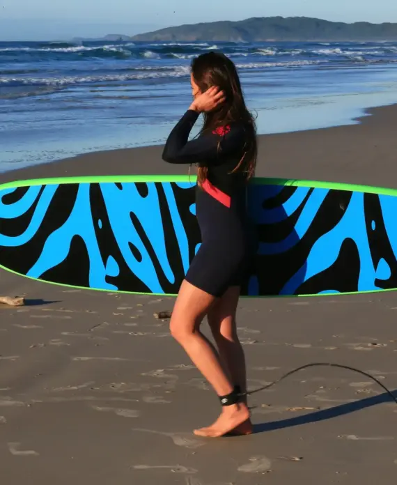 A person in a wetsuit walks barefoot on a sandy beach, carrying a surfboard with a bold blue and black pattern. Waves and distant hills are visible in the background.