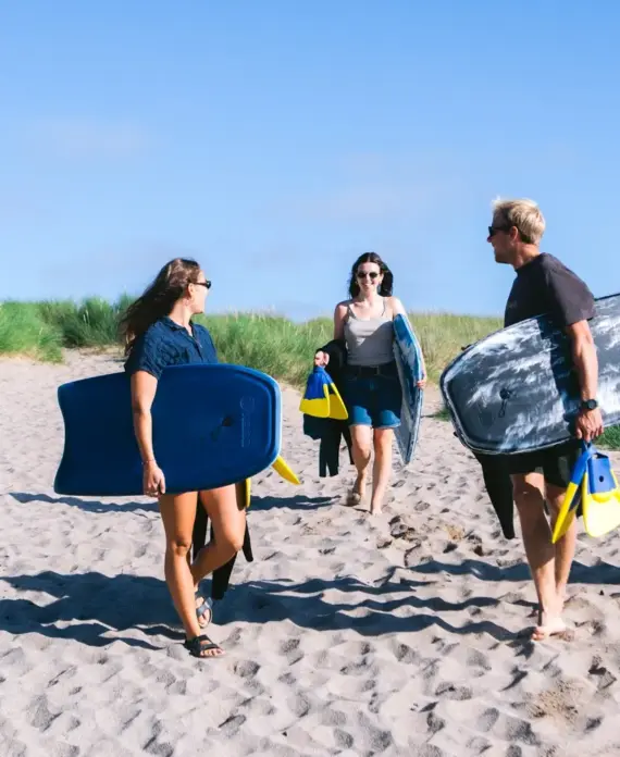 Four people walk on a sandy beach path with green grass, each carrying bodyboards and swim fins, enjoying a sunny day. Two adults walk ahead, while an adult and child follow behind. The sky is clear and blue.