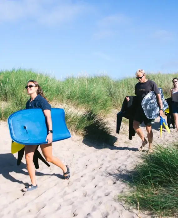 Four people walk on a sandy path through grassy dunes, carrying bodyboards and wetsuits on a sunny day, heading towards the beach.