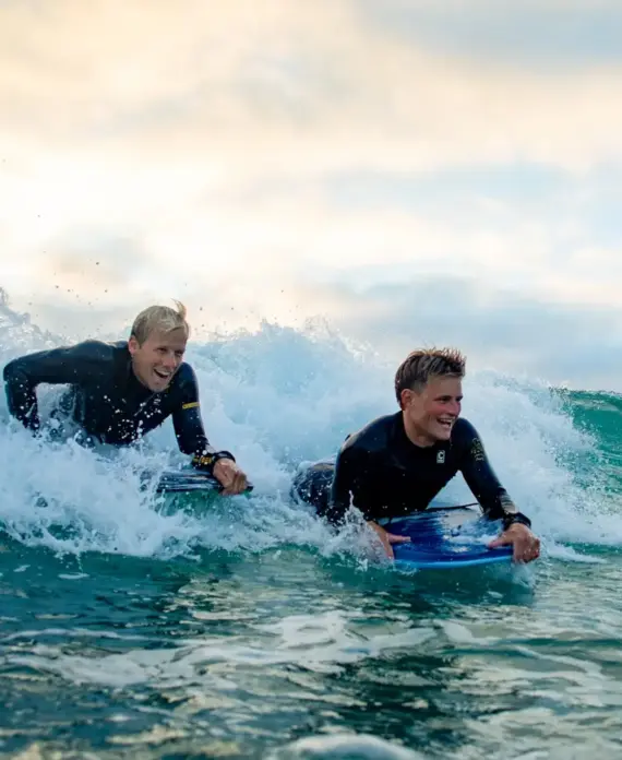 Two people in wetsuits ride bodyboards on an ocean wave, smiling and enjoying the water as the sun sets behind them, creating a bright and lively atmosphere.