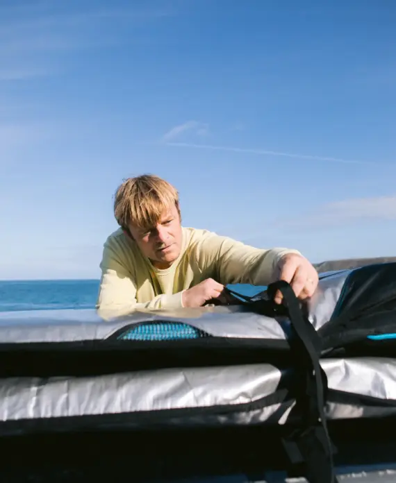 A person with blonde hair, wearing a light yellow shirt, fastens straps on top of silver and black equipment, with the sea and blue sky in the background.
