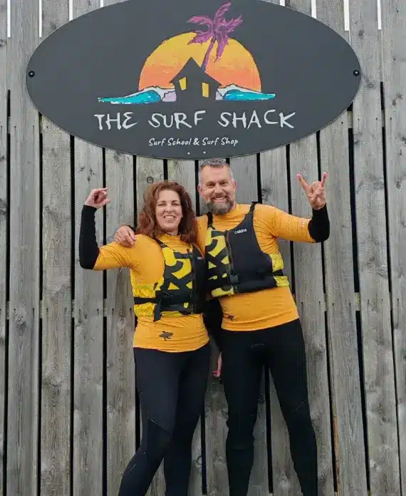 Two people in yellow life jackets and wetsuits stand smiling with arms raised in front of a wooden fence, beneath a sign reading “The Surf Shack” with a sunset and palm tree graphic.