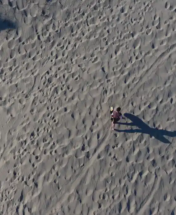 A person stands alone on sandy ground covered in footprints, casting a long shadow. The photo is taken from above, showing the textured sand and the solitary figure near the centre of the image.
