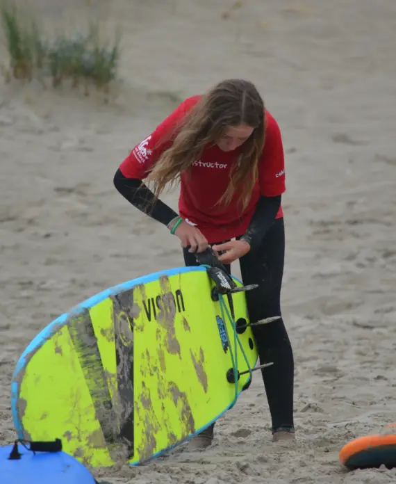 A person wearing a red instructor shirt and black wetsuit adjusts the fins on a bright yellow surfboard on a sandy beach. Other surfboards and dune grass are visible in the background.