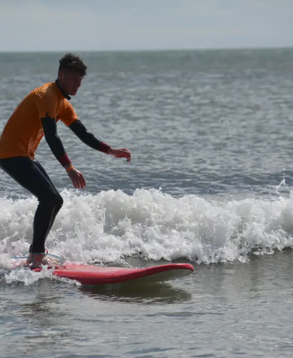A person wearing a wetsuit and an orange shirt is surfing on a small wave near the shore, standing on a red surfboard with the sea and horizon in the background.