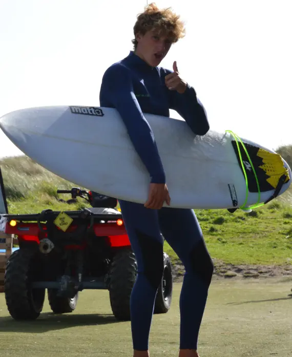 A person in a blue wetsuit stands outdoors holding a surfboard under one arm and giving a thumbs-up. A quad bike and grassy dunes are visible in the background.