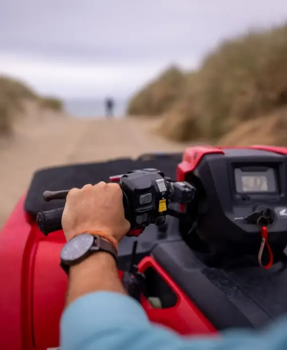 A person wearing a watch grips the handlebar of a red quad bike, driving on a sandy track between tall grass, with a blurred figure visible in the distance.