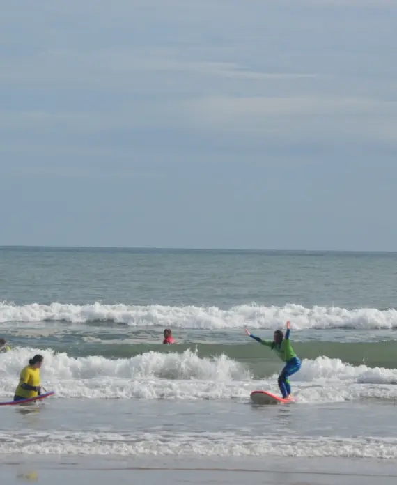 Several people wearing wetsuits and life jackets are surfing and playing in the shallow waves at a beach. Two individuals are riding surfboards near the shore, whilst others are in the water. The sky is cloudy.