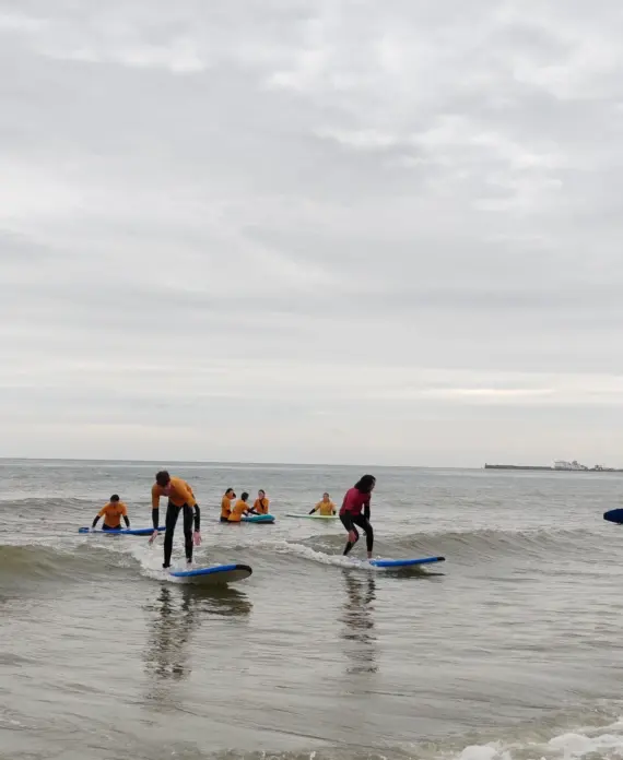 A group of people in wetsuits are surfing and paddling on surfboards in the sea near the shore under a cloudy sky. Some are standing on their boards whilst others are sitting or lying down.
