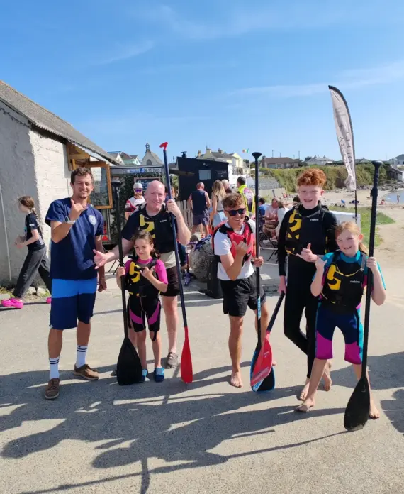 A group of six people, including children and adults in wetsuits, smile and hold paddles outside near a beach on a sunny day. Some give thumbs up, and a beach with people is visible in the background.