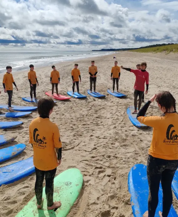 A group of children in yellow "Surf Shack" shirts stand on a sandy beach in a semicircle with surfboards, listening to two instructors giving instructions under a cloudy sky.