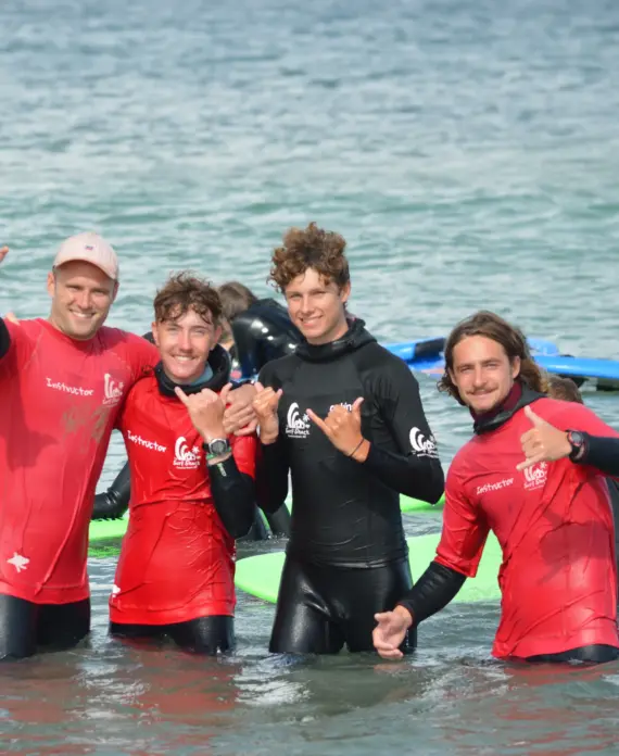 Four people in wetsuits and red instructor shirts stand in shallow water at the beach, smiling and making hand gestures, with surfboards and the sea visible in the background.