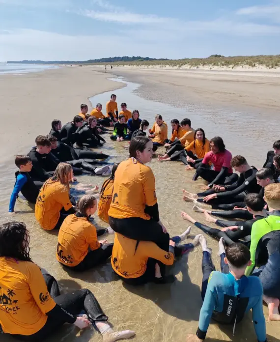 A large group of people in wetsuits, most wearing yellow shirts, sit in a circle on wet sand at the beach, listening to someone in the centre. The sky is clear and the beach stretches into the distance.
