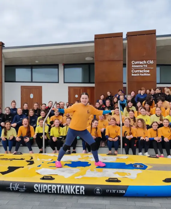 A large group of smiling children and adults in yellow and black shirts pose outside a modern building. One girl stands proudly at the front on a giant paddleboard labelled "SUPERTANKER.