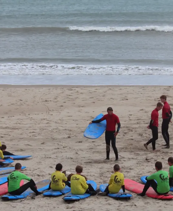 A group of people wearing wetsuits and coloured vests sit on surfboards on the sandy beach, listening to instructors in red shirts, with waves visible in the background.
