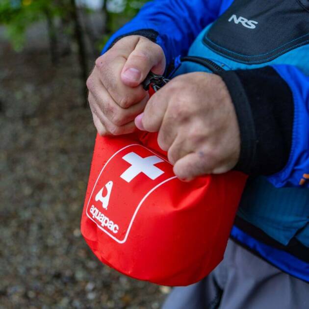A person in outdoor gear closes an Aquapac Waterproof First Aid Kit Bag with a white cross symbol, standing on a gravel surface.