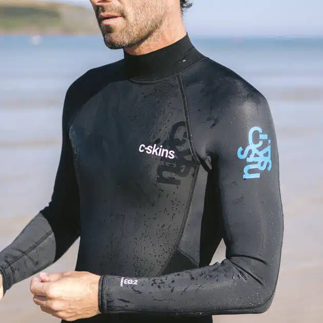 A man in a C-Skins Men's Element 3/2mm Back Zip Wetsuit stands on a sandy beach, with wet hair and the sea and distant hills behind him, appearing ready for water activities.