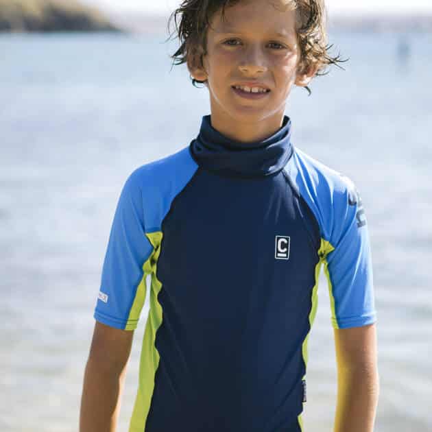 A young boy with wet hair stands on a beach under a clear sky, wearing the C-Skins Rash X Junior Short Sleeved Vest in blue and yellow, along with grey shorts. The sea and distant shoreline are visible in the background.