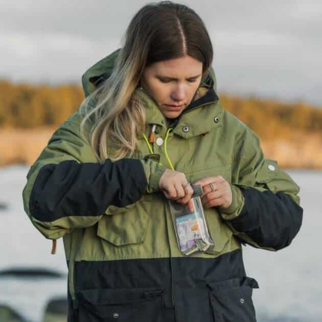 A woman in a green and black winter jacket stands outdoors by blurred trees and water, focusing as she places or removes items from an Aquapac Waterproof Keymaster with Lanyard.