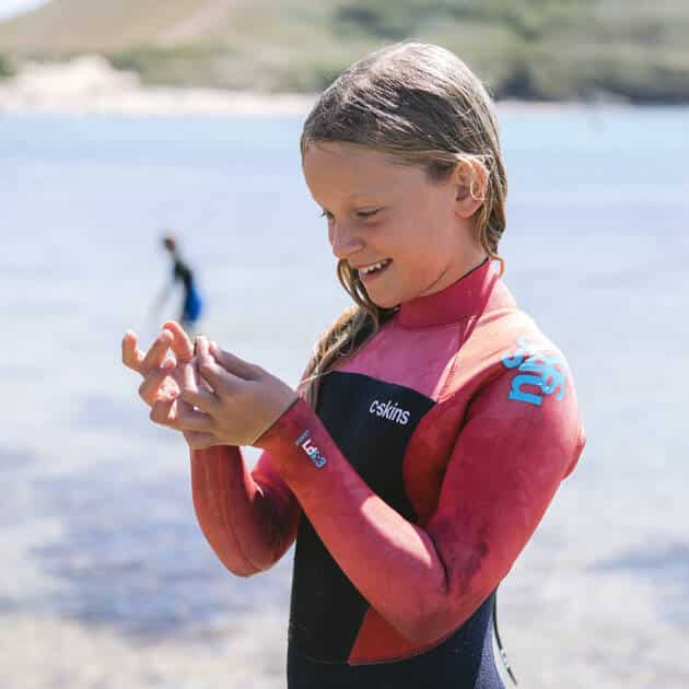 A young girl wearing the C-Skins Junior Legend 4/3mm Back Zip Wetsuit smiles as she examines something in her hands, standing in shallow, clear water with a blurred figure and hills visible in the background.