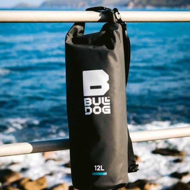 A Bulldog Dry Bag (12L, black) hangs on a metal railing with the sea, rocky shore, and distant cliffs beneath a blue sky.