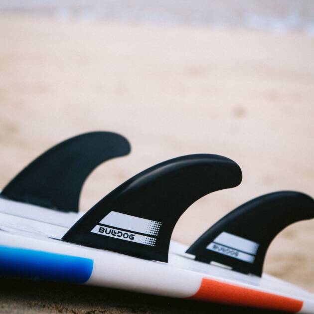 Close-up of Bulldog Moulded Thruster Twin Tab Fins (set of 3) on a surfboard with blue and red edges, resting on sandy beach with the ocean and sky softly blurred in the background.