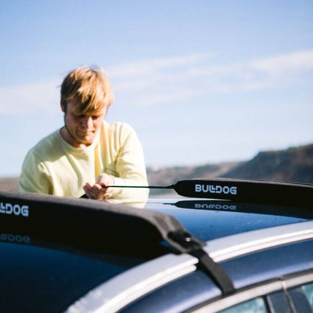 A person with blond hair secures gear to a Bulldog Single Soft Rack on a car outdoors on a sunny day, with hills and blue sky in the background.