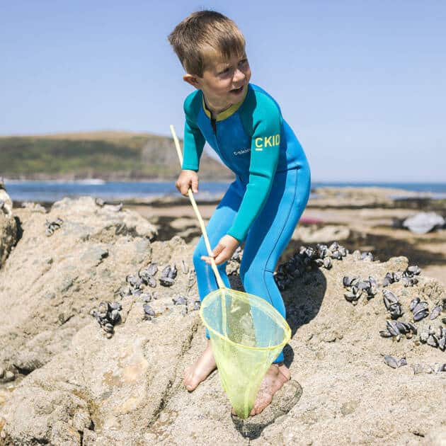 A young boy in a C-Kid Baby Full Length Wetsuit stands barefoot on rocky shore, holding a yellow-handled green net, gazing into a rockpool under clear blue sky with the sea and distant hills in the background.